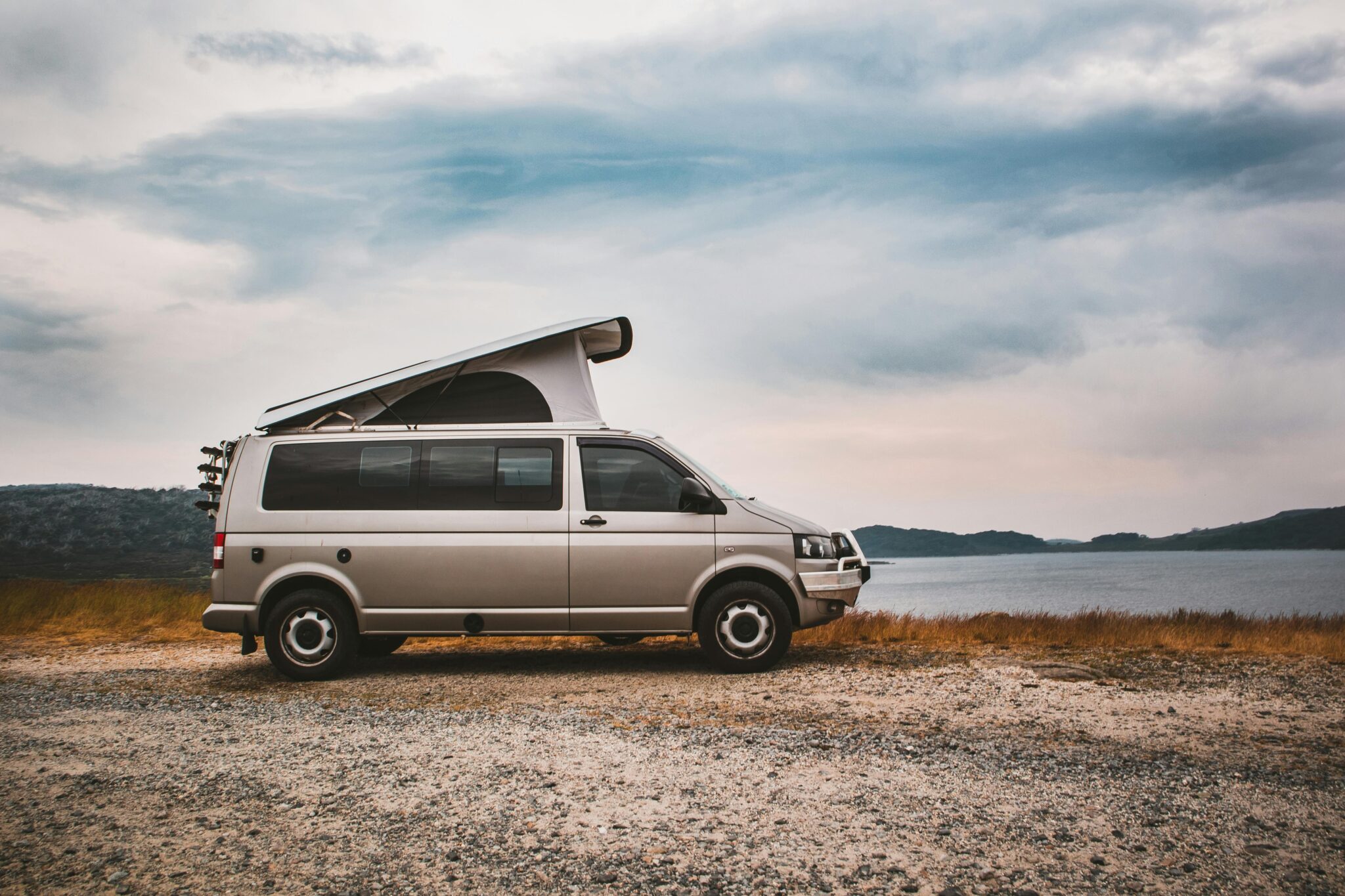 A camper van with rooftop tent parked by a tranquil lake under cloudy skies, ideal for adventure travel.